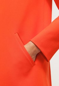 Detail jasně oranžového kabátu s skrytou boční kapsou a hladkým, strukturovaným materiálem s čistým švem na rukávu.