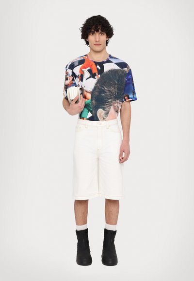 Shorts en denim blancs et un t-shirt graphique coloré présentant divers motifs. Le mannequin porte des bottes noires et tient un livre.