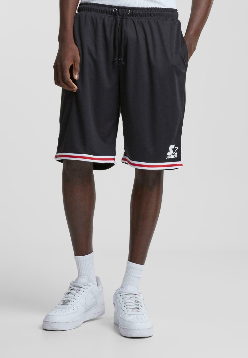 Bermudas deportivas negras con dobladillo a rayas rojas y blancas, cinturilla elástica y logo en la parte inferior izquierda. Combinadas con zapatillas blancas y calcetines.