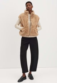 Hellbrauner Fake-Fur-Weste mit hohem Kragen und Frontreißverschluss, kombiniert mit einem beigen Oberteil und schwarzen, weiten Hosen. Einfaches, klares Design.