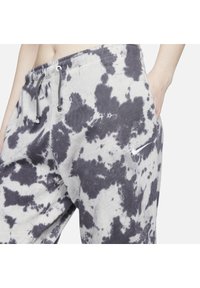 Osoba má na sobě šedé a černé tie-dye Nike tepláky s elastickým pasem a stahovacími šňůrkami, ruka v pravé kapse, viditelné bílé logo Nike.