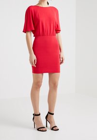 Kobieta w dopasowanej czerwonej sukience z falbanami na rękawach i czarnych sandałach na wysokim obcasie z paskiem wokół kostki na jednolitym białym tle.