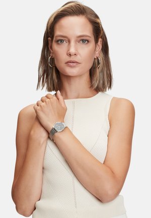 Zilveren polshorloge met een ronde wijzerplaat, dunne bezel en een gestructureerde metalen band. Model draagt een mouwloze, geribbelde crème top.