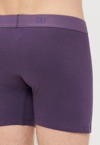 Boxers de algodão roxos com uma cintura elástica suave, apresentando o nome da marca "GAP" sutilmente em relevo. Textura suave e design ajustado.
