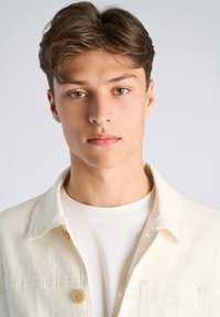 Jasna kurtka jeansowa na białej koszuli, z zapięciem na guziki i gładką fakturą. Modelka ma krótkie brązowe włosy, a tło jest neutralne.