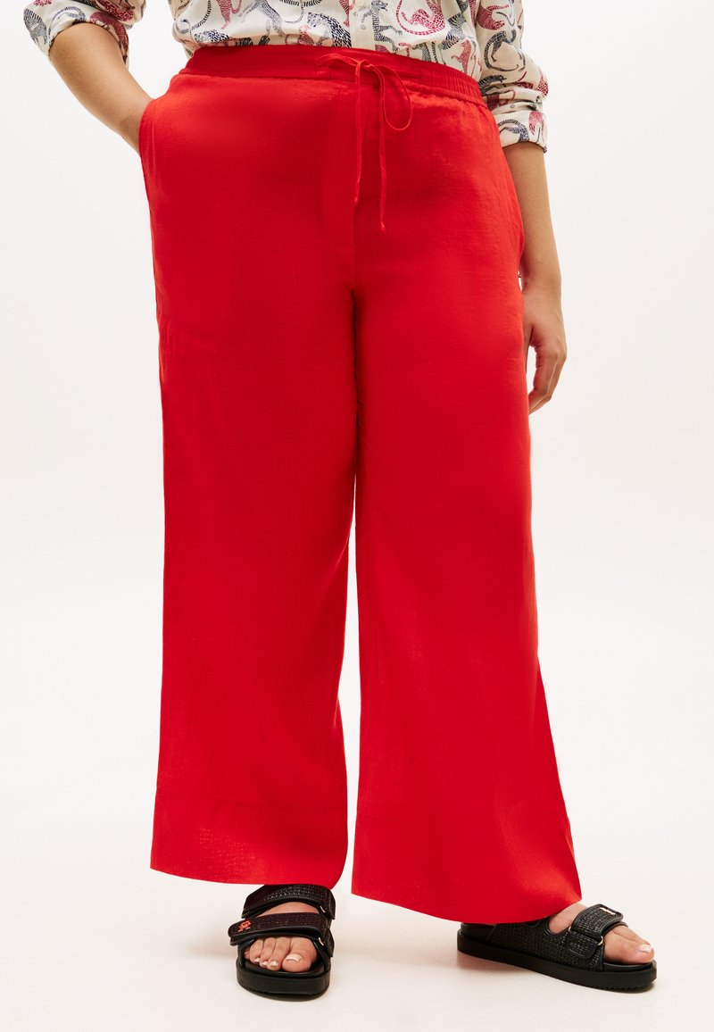 Κόκκινο παντελόνι με φαρδιά γραμμή, ελαστική μέση και κορδόνι. Το ύφασμα έχει λεία υφή. Συνδυασμένο με μαύρα σανδάλια με λουράκια.