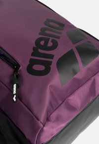 Zaino in tessuto viola con dettagli neri, caratterizzato da un logo e una cerniera. La superficie presenta una leggera texture con dettagli di cuciture visibili.