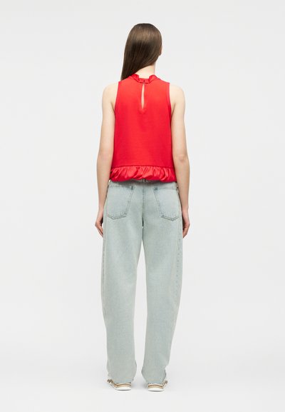 Γυναίκα με μακριά καστανά μαλλιά, φοράει κόκκινη αμάνικη μπλούζα με βολάν στο τελείωμα και ανοιχτό μπλε χαλαρά τζιν, στέκεται με την πλάτη στραμμένη σε λευκό φόντο.