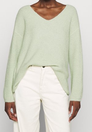 Personne portant un pull en maille vert clair à col en V, rentré dans un pantalon taille haute écru, debout les bras le long du corps.