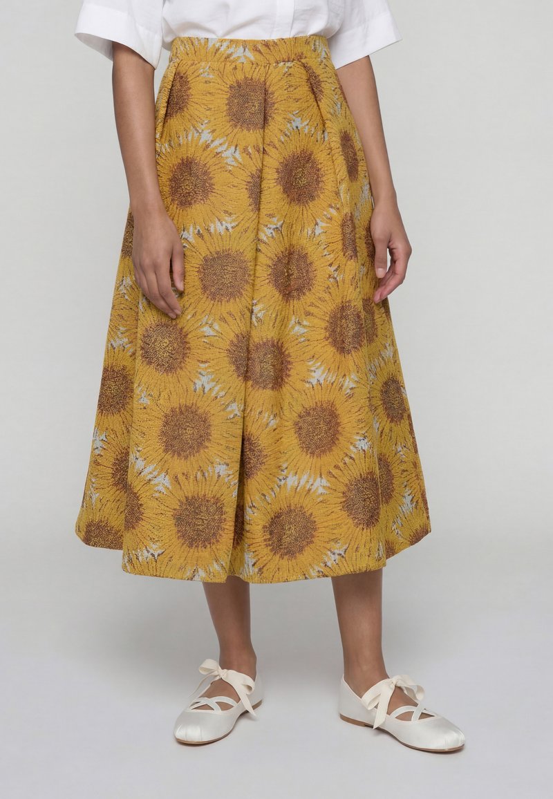 Personne portant une jupe midi jaune à motif tournesol, un t-shirt blanc à manches courtes et des ballerines blanches avec rubans à la cheville, debout face à l'avant.