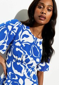 Mujer vestida con un vestido de estampado floral azul y blanco, con mangas cortas abullonadas, posando contra un fondo blanco liso.