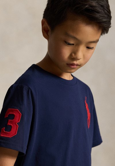 Noor poiss vaatab alla, kandes tumesinist särki, millel on suur punane number 3 varrukal ja punane tikitud muster rinnal.