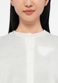 Cardigan tricoté blanc avec un col rond, des boutons sur le devant, et un subtil logo "Hugo" en forme de cœur sur le côté gauche.
