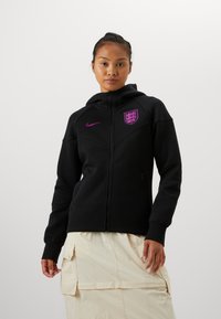Melns zip-up džemperis, kas izgatavots no gludas auduma, ar purpursarkanu Nike logo un Anglijas emblēmu. Ietver priekšējas kabatas un pieguļošas kapuces.