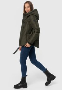 Green padded jacket with a hood, zippered front, side pockets, and black accents. Paired with blue jeans and black chunky ankle boots.