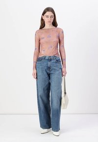 Langermet, gjennomsiktig topp i fersken med blå mønstre, kombinert med høytliv, løstsittende lyseblå jeans og hvite hæler. Holder en beigesekk.