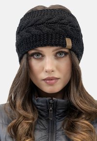 Bandeau en tricot noir avec un motif tressé texturé, arborant une petite étiquette en cuir beige. Porté avec une veste matelassée sombre.