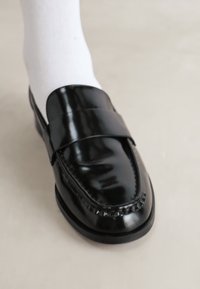 Svart skinnloafers med en glatt, blank overflate, med en bred stropp og detaljerte sømmer langs kantene, båret med hvite sokker.