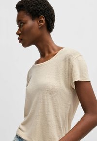 Camicia beige a maniche corte realizzata in tessuto leggero, con scollatura ampia e una delicata trama. Design semplice e informale senza motivi prominenti.