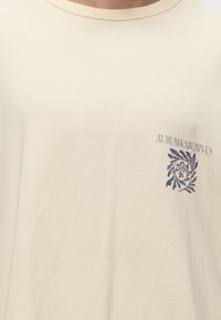 Μπεζ μπλουζάκι με στρογγυλή λαιμόκοψη με μικρό σκούρο μπλε λουλουδάτο σχέδιο και το κείμενο "AURUM&ARCHIVO" στο αριστερό στήθος.