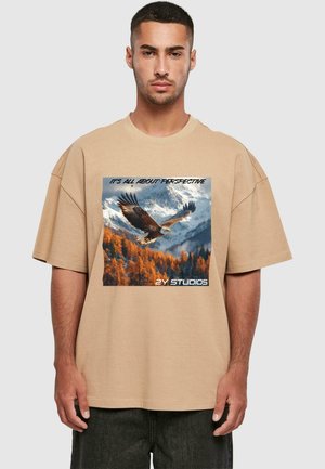 Beige oversized t-shirt featuring a printed eagle design against a mountain backdrop. Text reads "IT'S ALL ABOUT PERSPECTIVE" and "2Y STUDIOS."