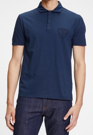Homme portant un polo bleu marine à manches courtes avec trois boutons et un logo discret sur la poitrine, associé à un jean bleu foncé.
