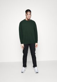 Zelená mikina s kulatým výstřihem a žebrovanými manžetami, doplněná o černé kalhoty a bílé sportovní boty. Pozadí je čistě bílé.