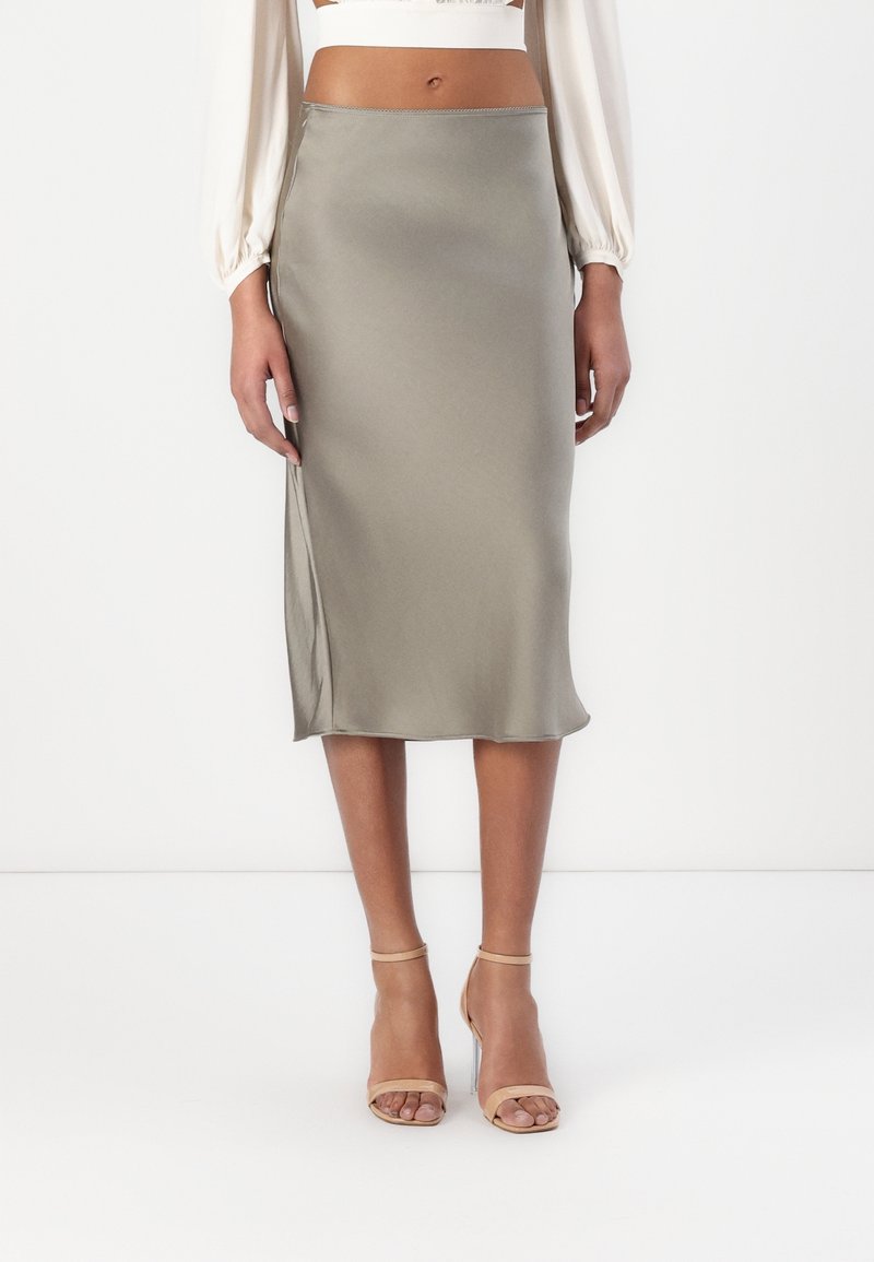 Femme portant une jupe en satin taupe taille haute jusqu'aux genoux, associée à des sandales à talons hauts nude à bride cheville, debout devant un fond uni.