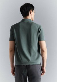 Hombre de espaldas que lleva una camisa polo verde de manga corta y pantalones oscuros sobre un fondo liso de color gris claro.