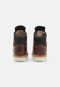 Botas de tornozelo em pele castanha com um colarinho acolchoado preto, sola de borracha bege e detalhe em costura laranja. Vista traseira mostrando as etiquetas de tamanho no interior.