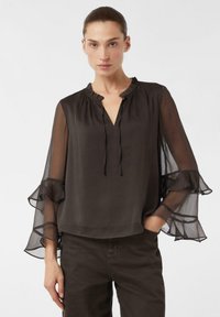 Blouse noire légère avec un col à volants et des manches longues transparentes agrémentées de détails froncés, associée à un pantalon sombre.