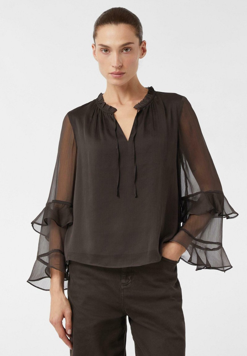 Blouse noire légère avec un col à volants et des manches longues transparentes agrémentées de détails froncés, associée à un pantalon sombre.