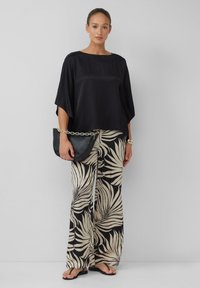 Haut en satin noir à manches larges, associé à un pantalon beige avec un motif de feuilles noires. Le modèle tient une pochette noire avec une chaîne dorée.