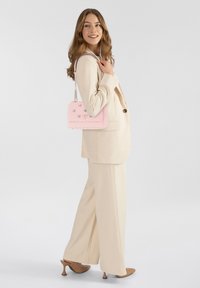 Borsa a tracolla imbottita rosa con motivi a diamante, tracolla a catena e logo in rilievo. Il modello indossa un completo crema e tacchi marroni. Sfondo semplice.