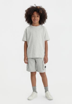 Barn med krøllet hår står og smiler iført lysegrå t-shirt, matchende cargoshorts, grå sokker og hvide sneakers på hvid baggrund.