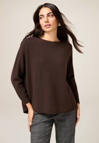 Brązowy sweter na drutach o luźnym kroju, z szerokim dekoltem i długimi rękawami, zestawiony z szarymi spodniami w kratę. Gładka tekstura, prosty design.