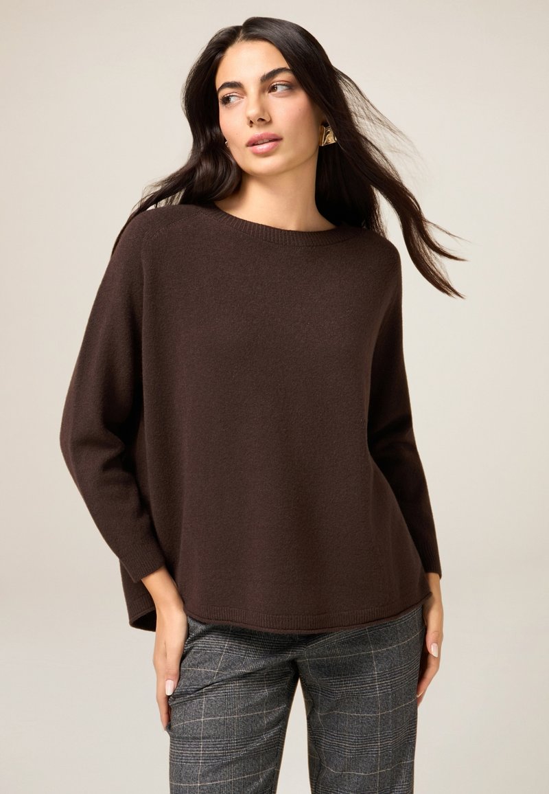 Brązowy sweter na drutach o luźnym kroju, z szerokim dekoltem i długimi rękawami, zestawiony z szarymi spodniami w kratę. Gładka tekstura, prosty design.