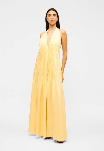 Γυναίκα που στέκεται, φοράει αμάνικο, κίτρινο μάξι φόρεμα με βαθύ V-λαιμόκοψη και πιέτες, με μαύρα ανοιχτά παπούτσια, πάνω σε απλό φωτεινό φόντο.