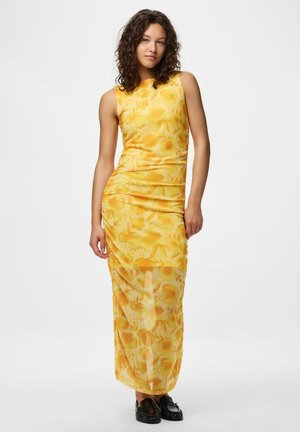 Femme aux cheveux bouclés portant une robe maxi jaune à fleurs sans manches et des mocassins noirs, debout devant un fond uni.