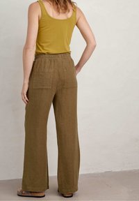 Žena v olivově zelených širokých kalhotách s zadními kapsami, hořčicovém topu bez rukávů a hnědých sandálech, stojící proti světlé zdi.