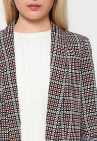 Il blazer a quadri presenta tonalità di marrone, blu navy e verde, con rever strutturati e una blusa bianca testurizzata sotto.