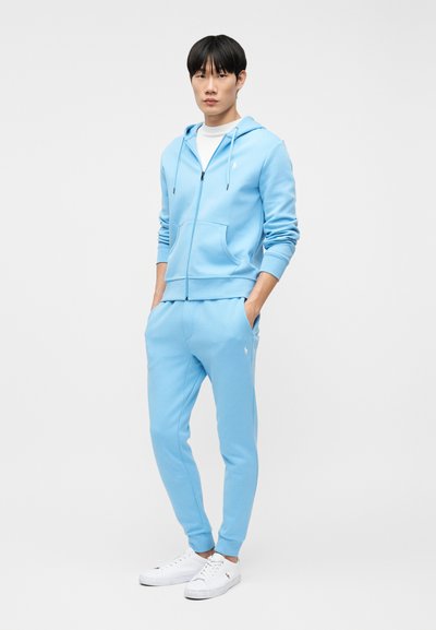 Νέος άνδρας που στέκεται, φορώντας ανοιχτό γαλάζιο φούτερ με φερμουάρ και ταιριαστό παντελόνι φόρμας, λευκά αθλητικά παπούτσια, με τα χέρια στις τσέπες, ουδέτερο φόντο.