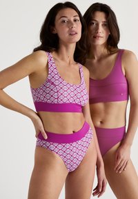 Zwei Frauen tragen passende Loungewear: Eine in einem gemusterten, pinken Bralette und hochgeschnittenen Unterhosen, die andere in einem einfarbigen, pinken Bralette und Unterhosen.