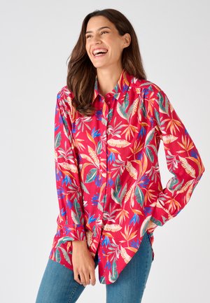 Mujer sonriendo con una camisa roja brillante de manga larga con botones y patrones de hojas tropicales multicolores y jeans azules, de pie frente a un fondo liso.