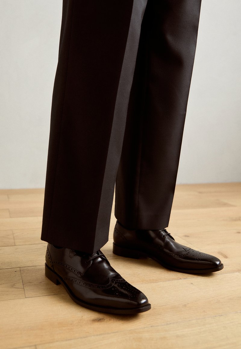 Schwarze Leder-Oxfords mit Wingtips-Design, versehen mit Brogue-Verzierungen und einem glänzenden Finish, kombiniert mit maßgeschneiderten schwarzen Hosen.