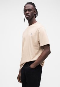 Beżowa bawełniana koszulka z krótkim rękawem, z małym niebieskim haftem "II" na piersi. Noszona z czarnymi jeansami.