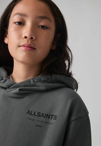 Felpa grigia con cappuccio e cordoncino, dotata di tasca frontale. Logo "ALLSAINTS" in nero sul lato sinistro, tessuto testa di moro, vestibilità leggermente ampia.