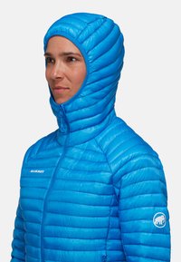 Blå isolerad jacka med hög krage och åtsittande huva, med horisontellt quiltade sektioner och en logo på ärmen. Dragkedjestängning fram.