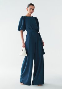 Turkoois jumpsuit met opgeblazen mouwen, een taille met ceintuur en wijde pijpen. Gecombineerd met een kleine witte handtas en sandalen met gouden hakken. Soepele stoftextuur.