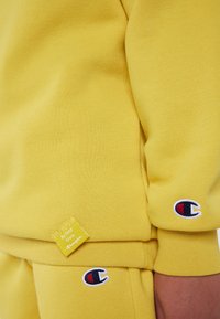 Sweat-shirt et pantalon de jogging jaunes avec des écussons du logo Champion sur la manche et la jambe, et une étiquette "Be Your Own Champion" sur l'ourlet du sweat-shirt.